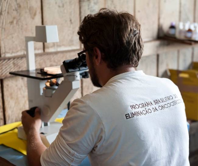 Técnico olhando lâmina ao microscópio usa a camiseta do programa brasileiro de eliminação da oncocercose